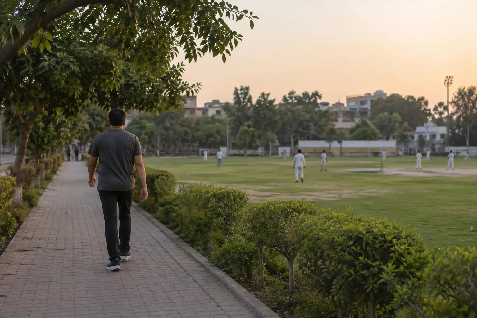A man walking along a path beside a neighbourhood cricket ground in soft evening light.