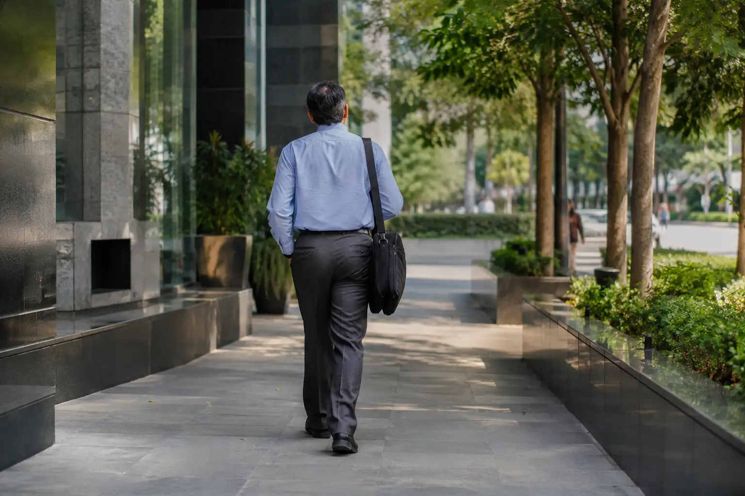 Man in office clothes and walking shoes walking along a shaded path beside an office building.
